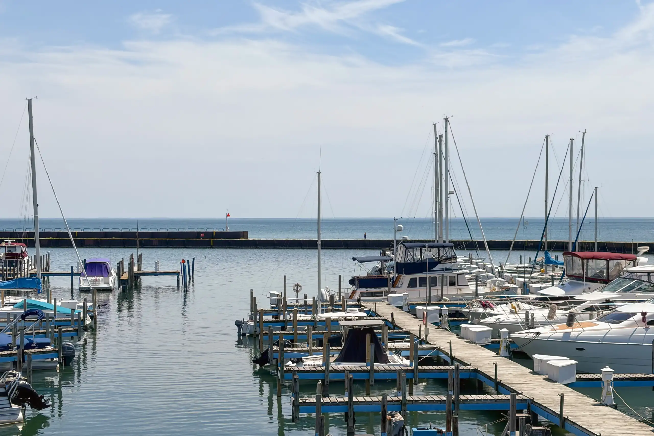 A wide shot of a marina filled with docked sailboats and powerboats, featuring wooden piers and a distant breakwater on a sunny day.
