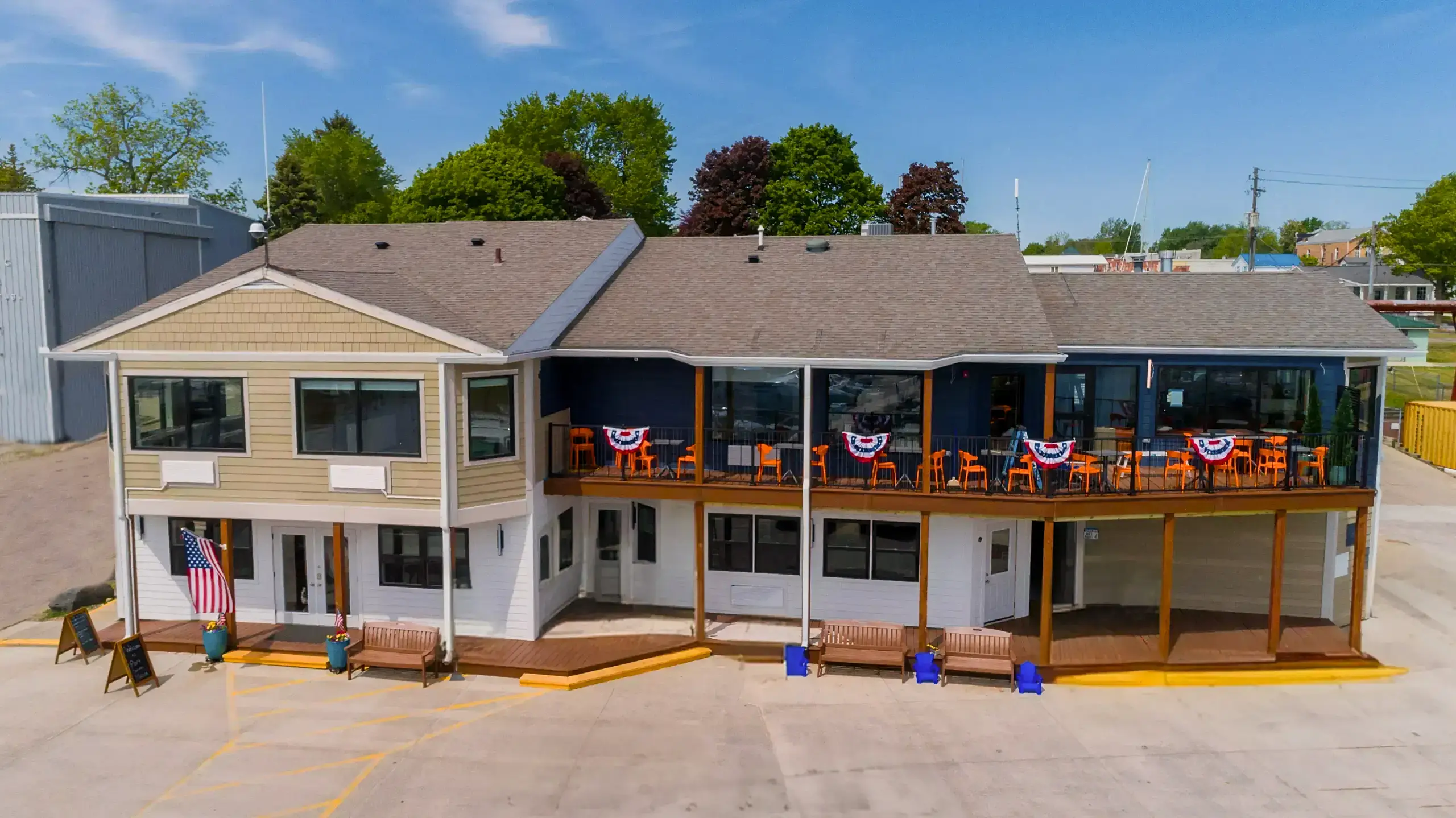 An aerial view of a two-story waterfront building, likely a restaurant, with tan siding and a dark blue accent. The upper level features a dining deck with orange chairs and patriotic bunting.