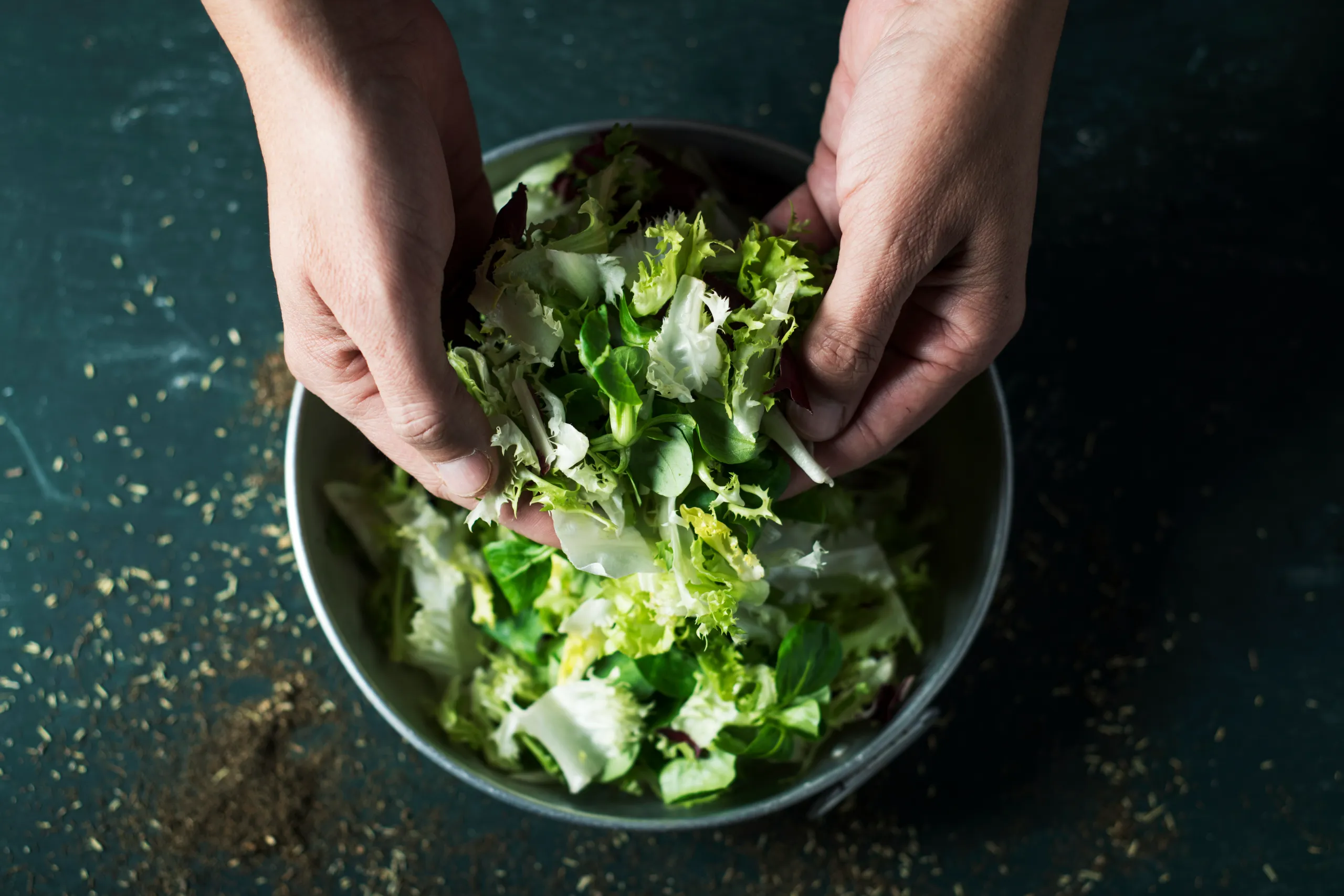 Two hands are seen from above, tossing and mixing fresh green and red lettuce leaves in a silver bowl, set against a dark counter.