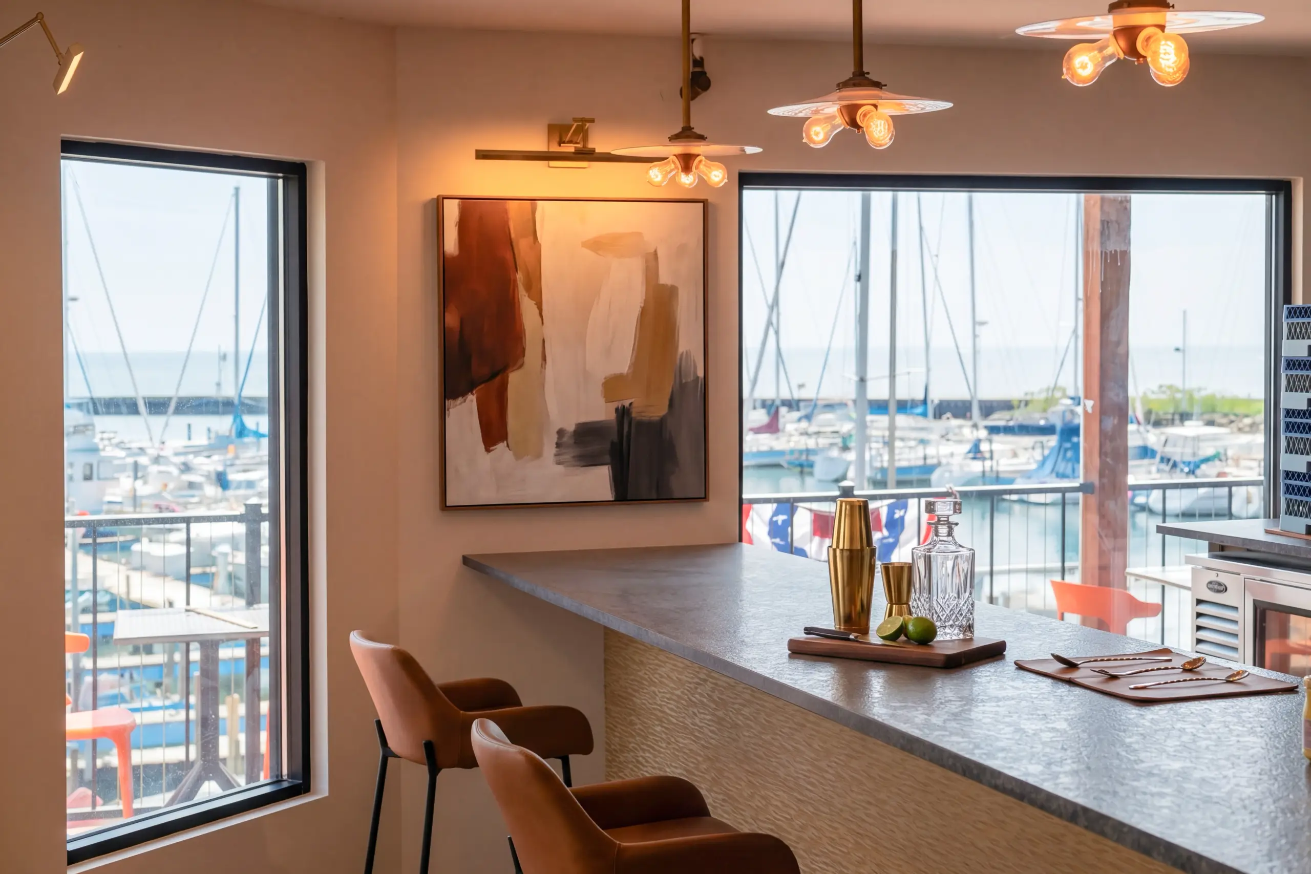 A stylish bar counter with two leather stools overlooking a sunny marina filled with boats. Cocktail tools and a decanter rest on the gray countertop.