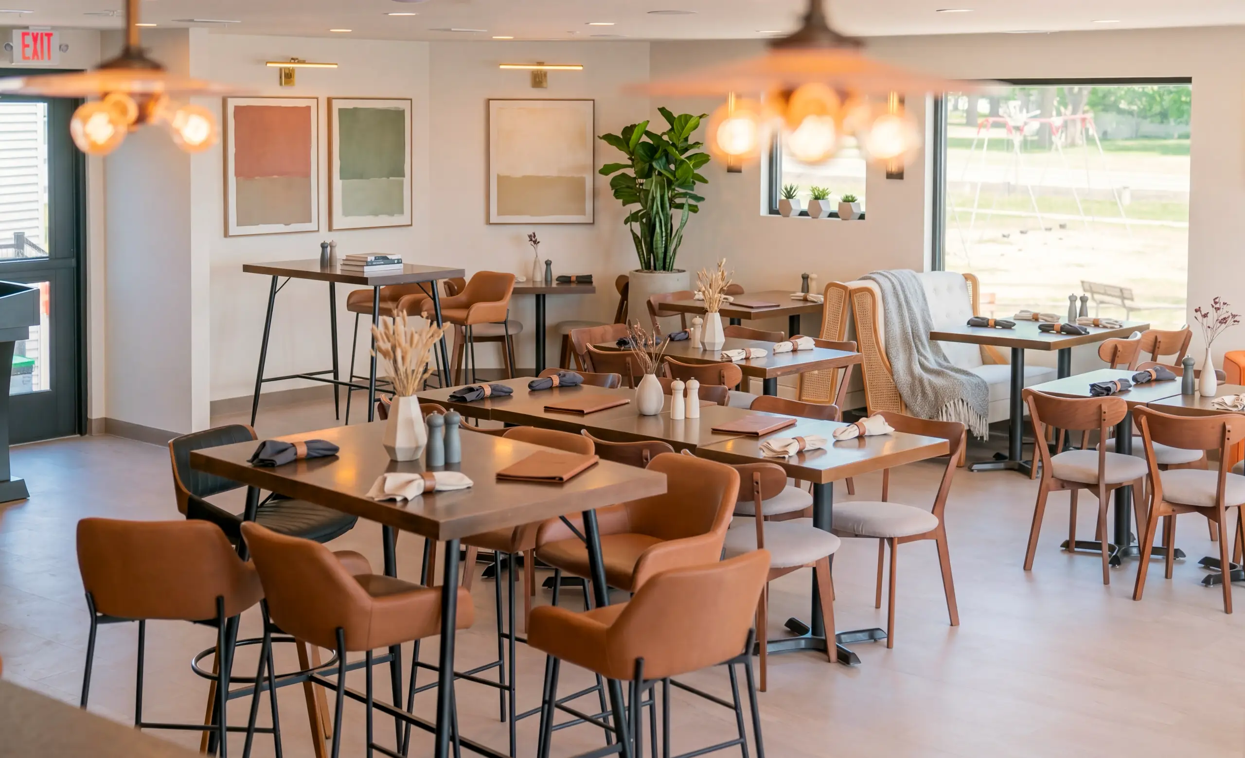 A modern, airy restaurant dining area with dark wooden tables set for diners. The room features brown leather chairs, abstract wall art, and large windows bringing in natural light.