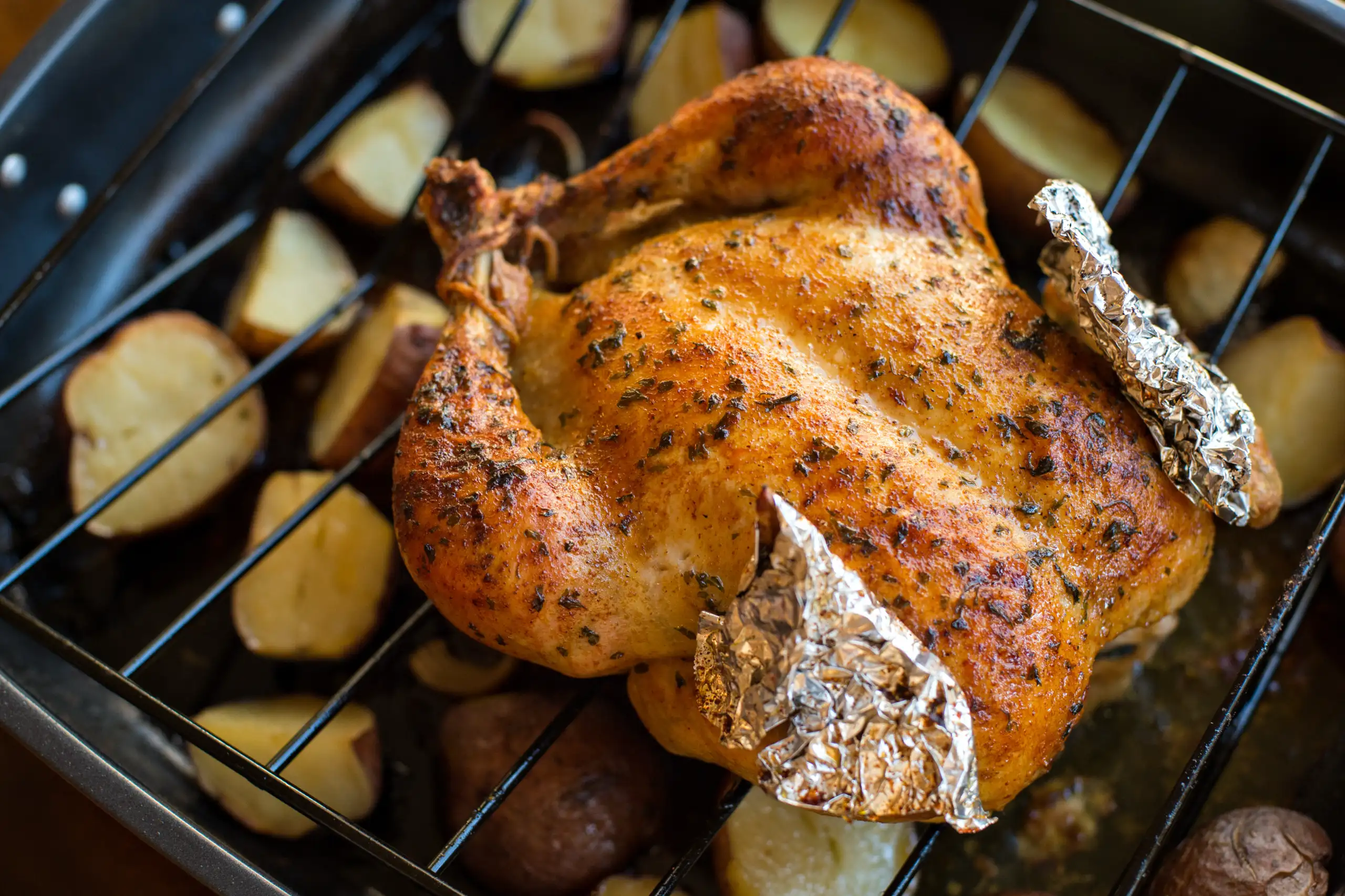 A whole roasted chicken with crispy, herb-seasoned skin sits on a rack in a roasting pan filled with halved potatoes.