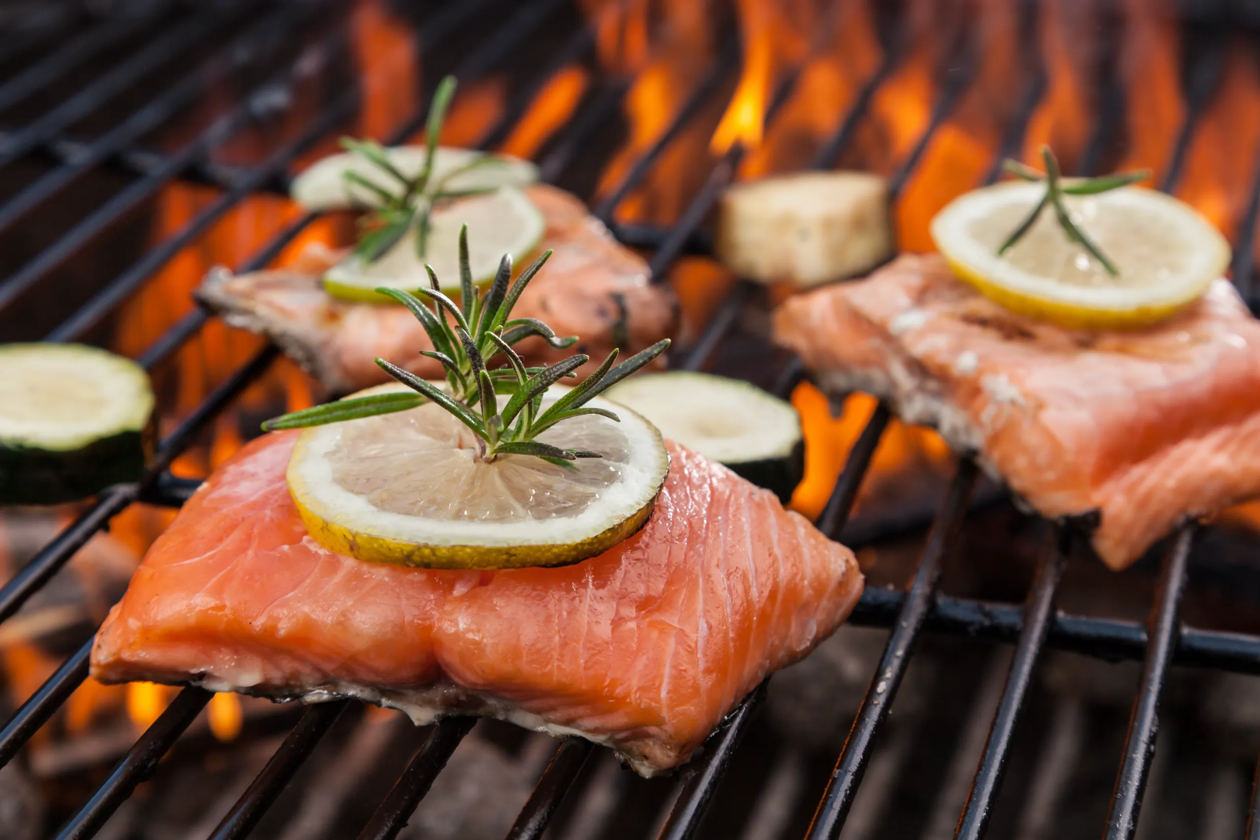 Fresh salmon fillets topped with lemon and rosemary sprigs sizzle on a barbecue grill over intense open flames, with slices of zucchini also cooking.