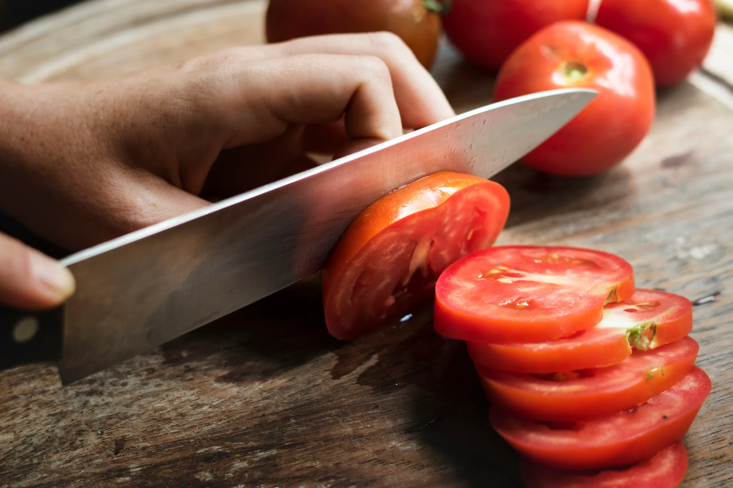 A hand uses a large chef's knife to slice a fresh, red tomato on a dark wooden cutting board. Several whole tomatoes and stacked slices are visible.