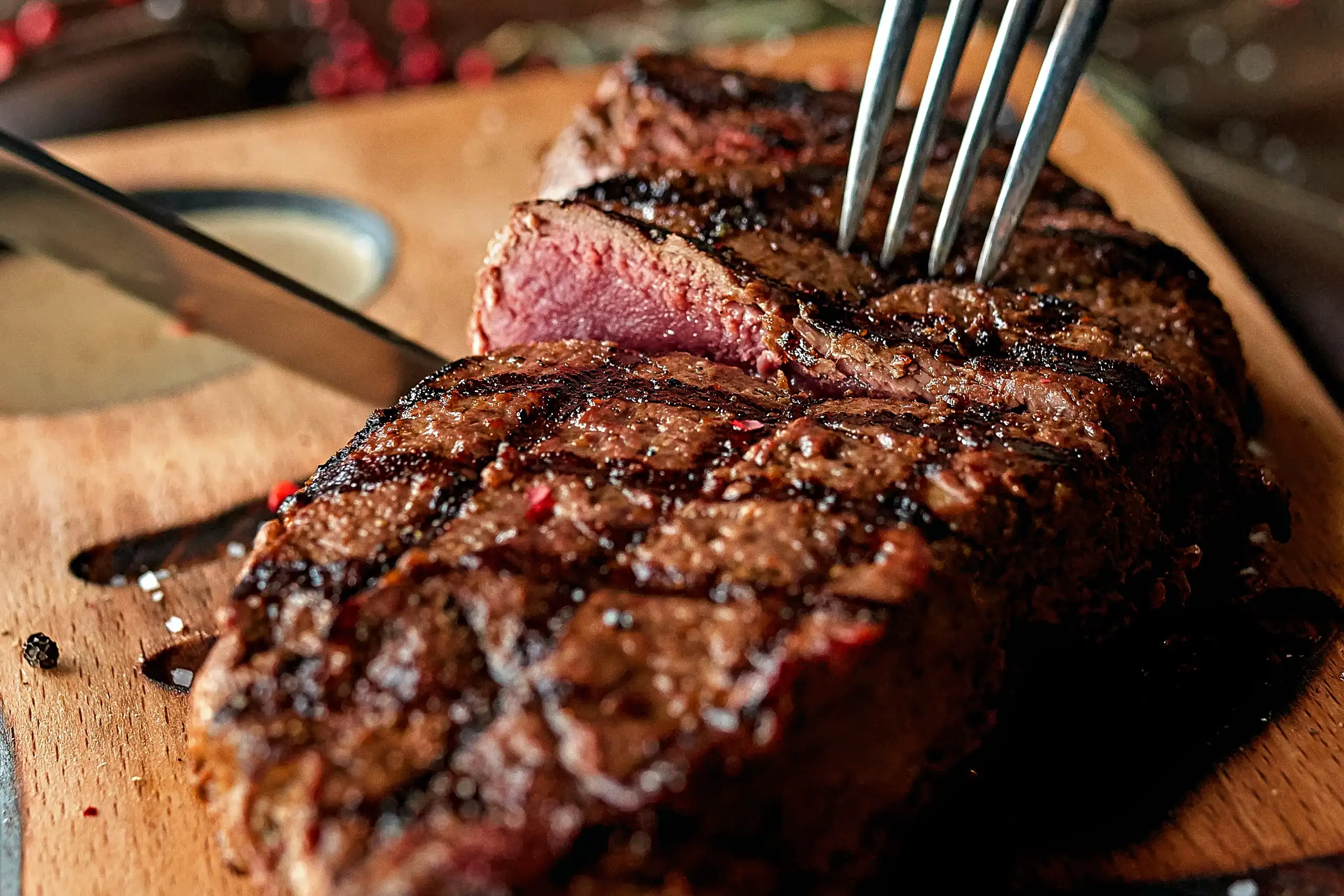 A close-up of a perfectly grilled, medium-rare steak being held by a fork on a wooden cutting board next to a knife.