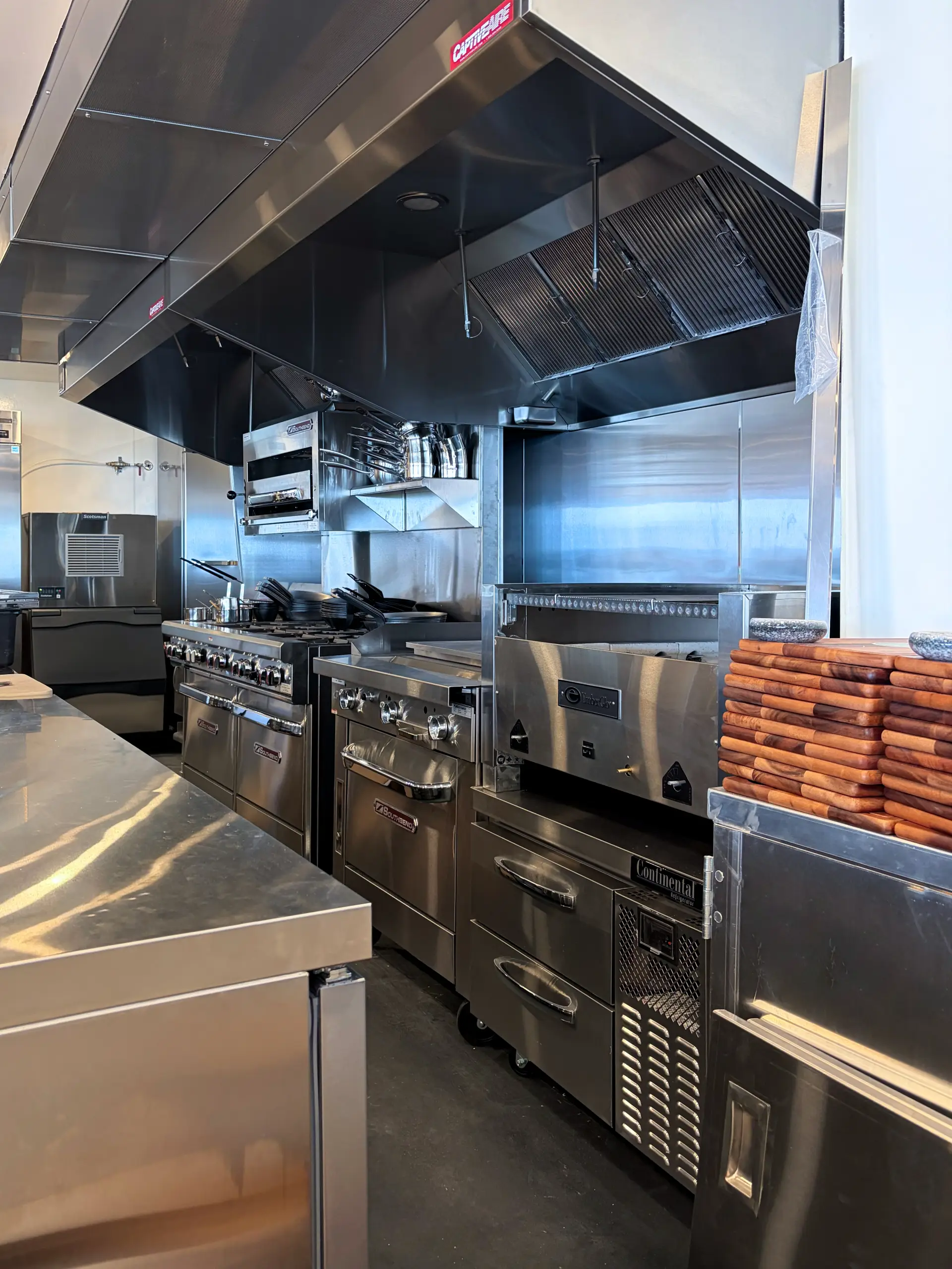 A professional commercial kitchen dominated by shiny stainless steel appliances, including an industrial stove, griddle, and large overhead vent hood.