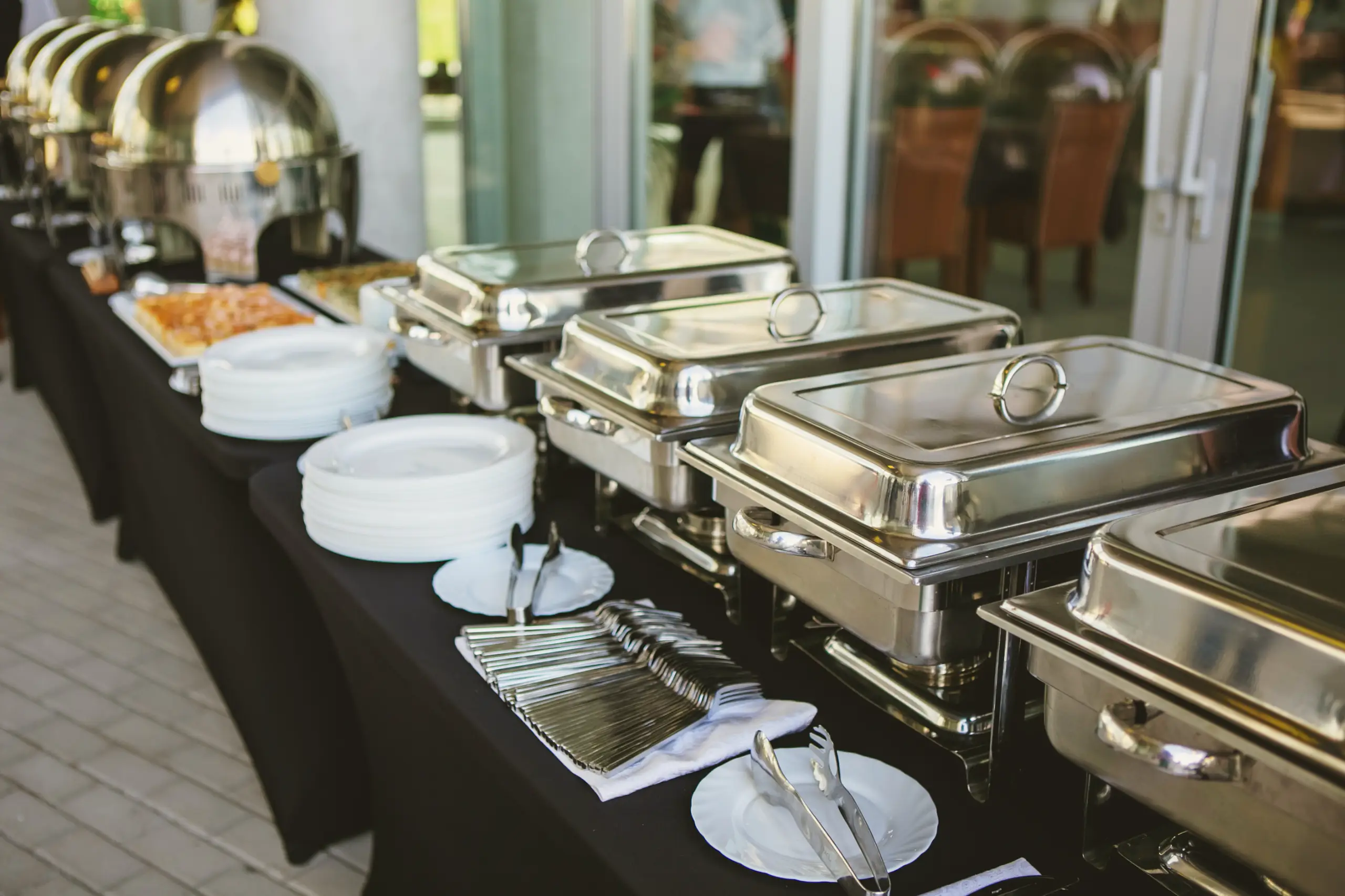 A professional buffet table with a black cloth features several silver chafing dishes, stacks of white plates, and organized silverware for a catered event.