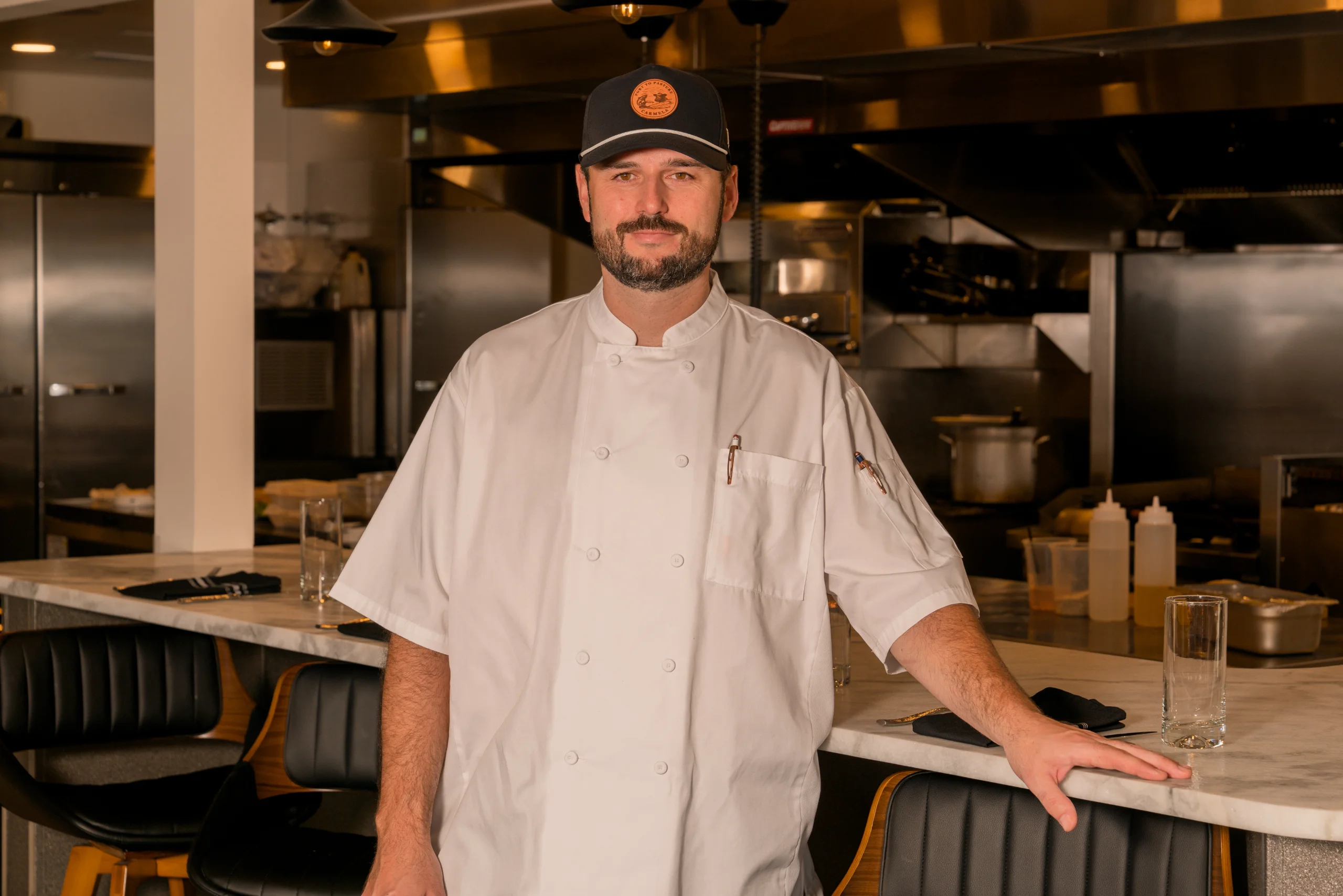 Chef Clint Frenette, in a white chef's coat and dark cap, stands leaning on a marble counter overlooking a professional stainless steel kitchen.