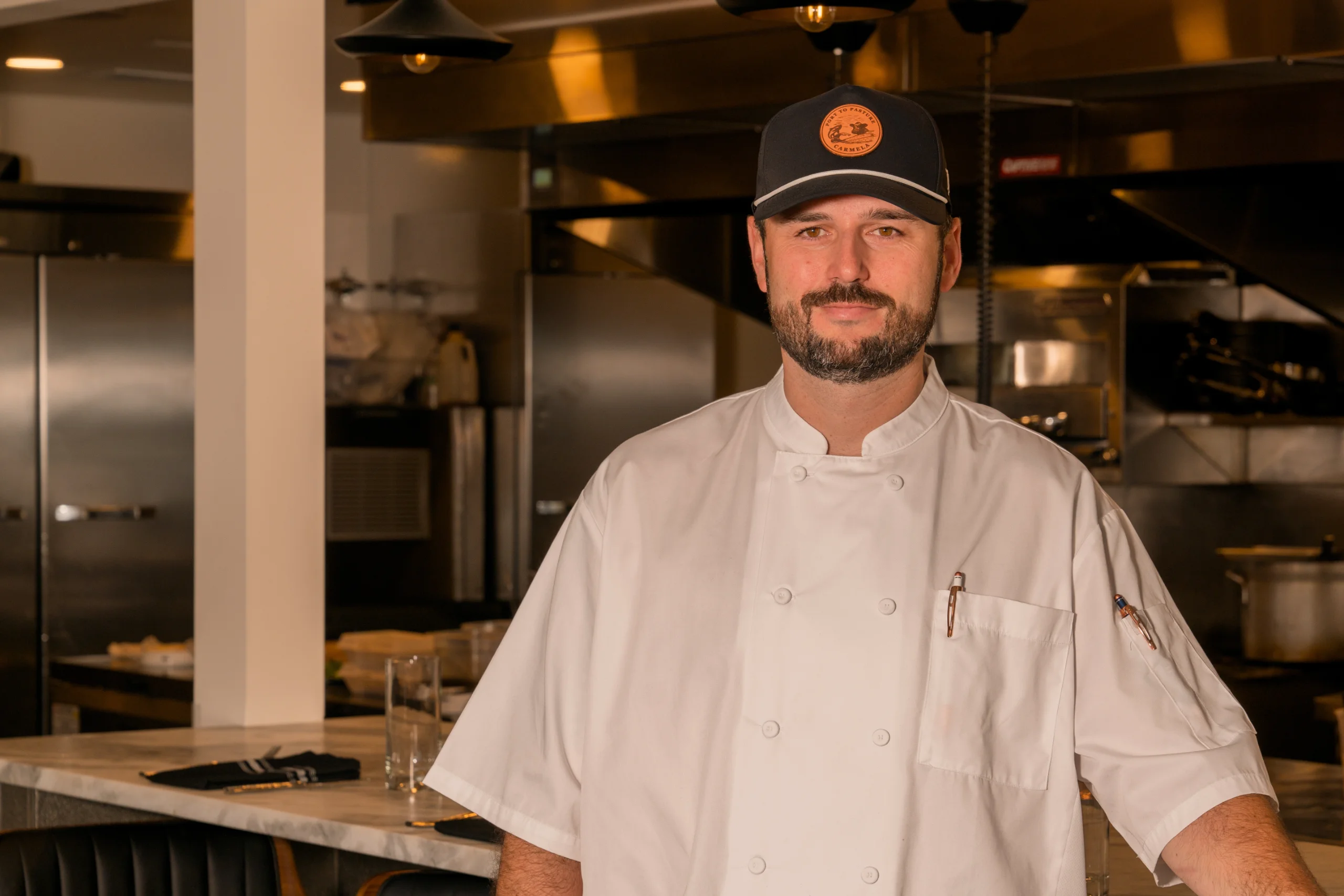 Chef Clint, wearing a white coat and dark baseball cap, stands in a professional restaurant kitchen environment, looking confidently at the camera.