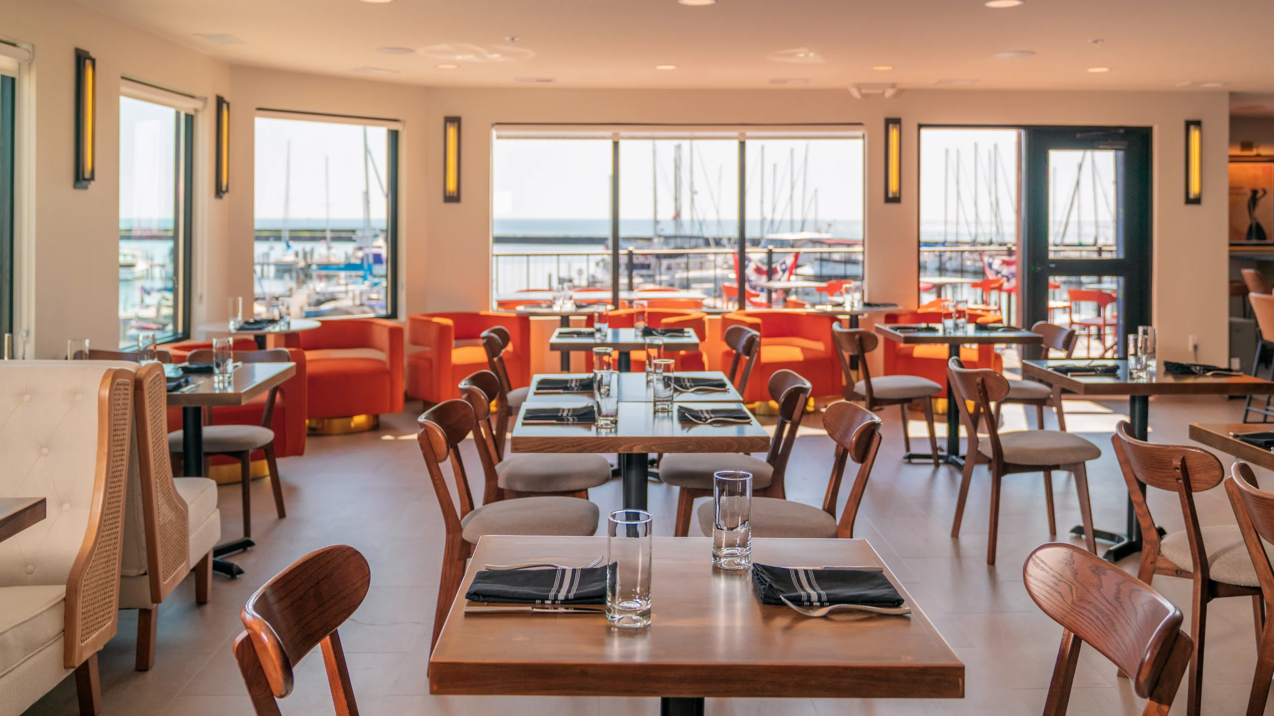 A modern restaurant dining room featuring set wood tables, beige booths, and bright orange seating, overlooking a sunny marina filled with docked boats.