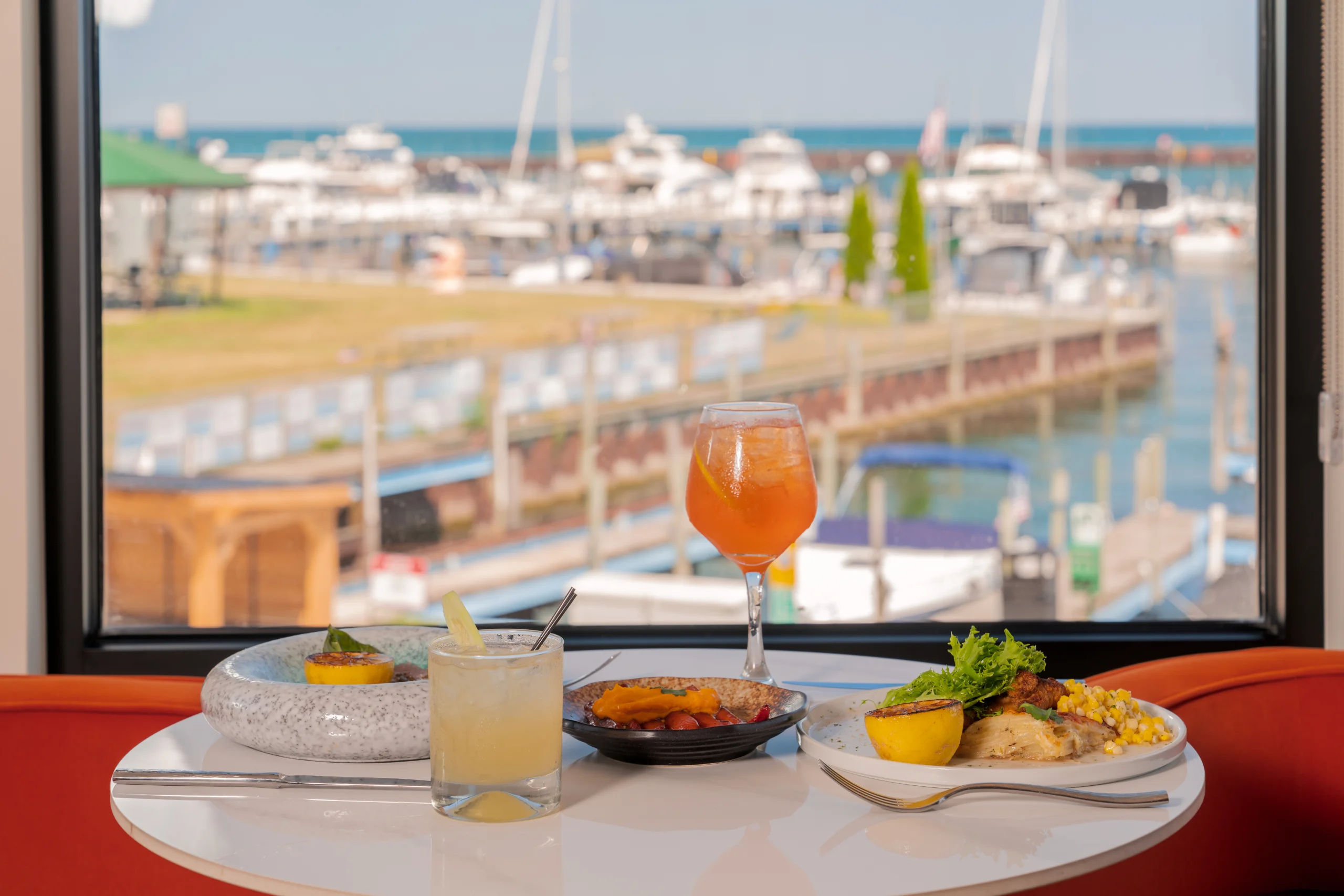 Two cocktails and several plates of food are arranged on a white table in front of a window overlooking a sunny marina filled with white yachts.