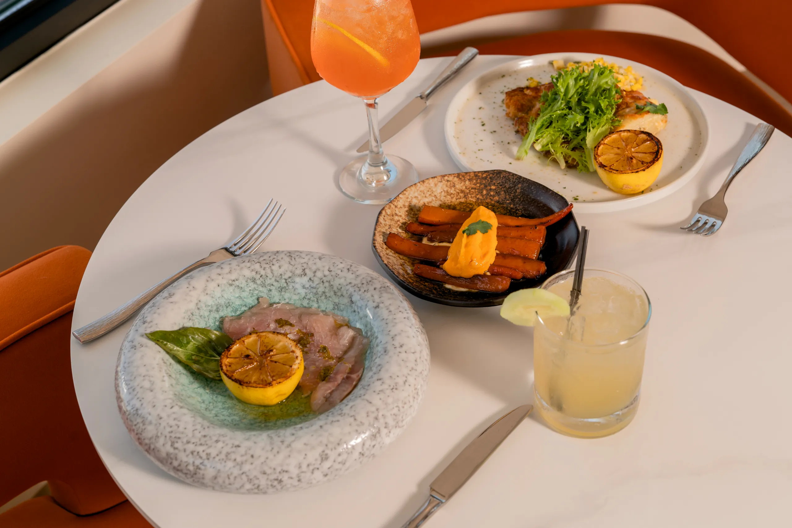 A white restaurant table set for two features three colorful, plated gourmet courses: fish crudo, glazed carrots, and a protein with salad, next to two cocktails.
