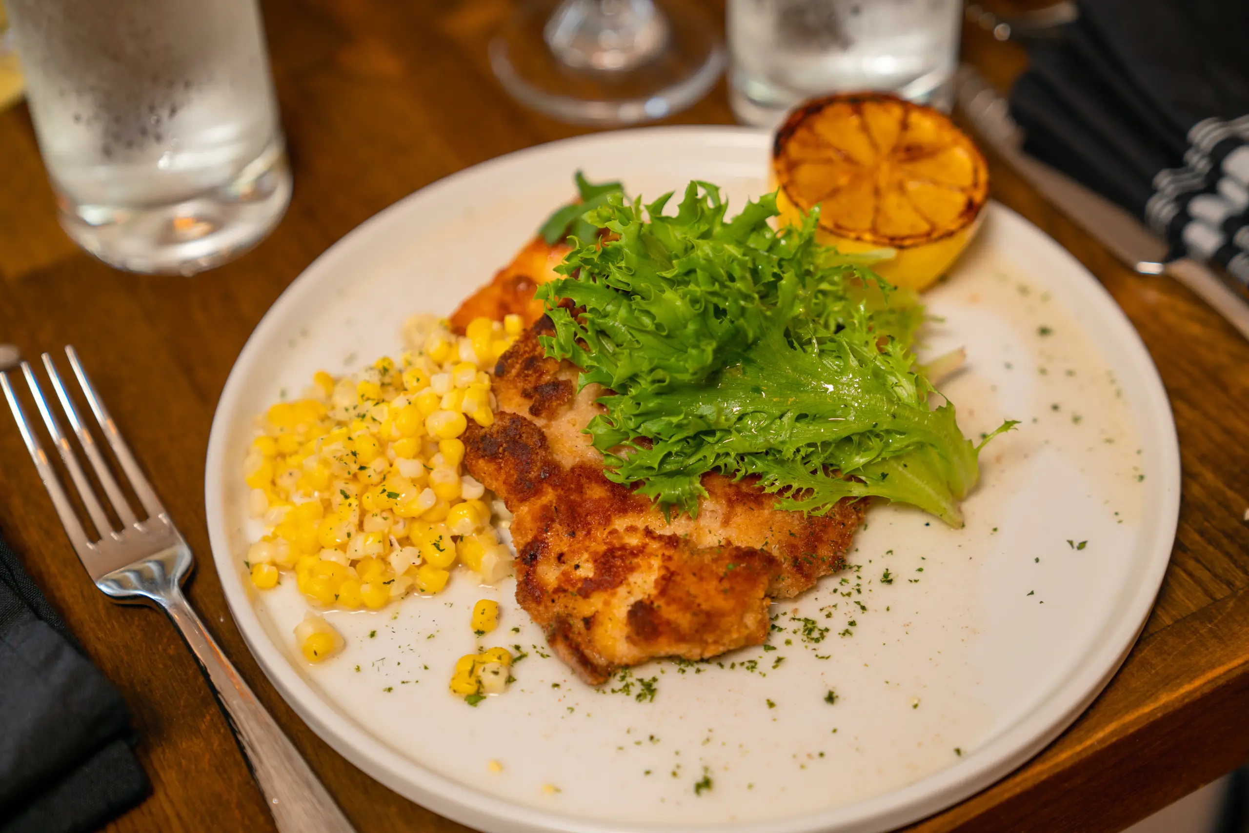 A crispy chicken cutlet is served with yellow corn, fresh lettuce, and a charred lemon wedge on a dinner plate.