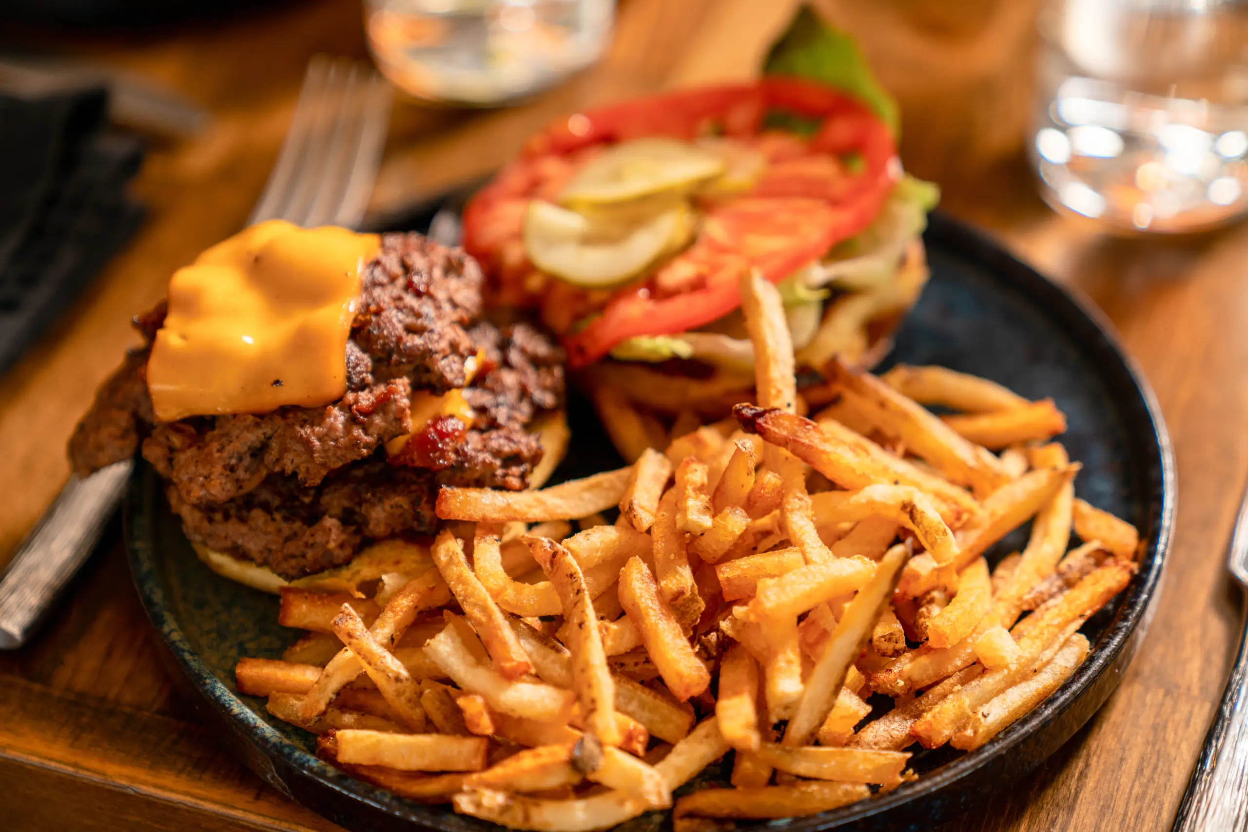 A dark plate holds a double smash burger topped with melted cheese, served next to a large pile of golden, crispy french fries.