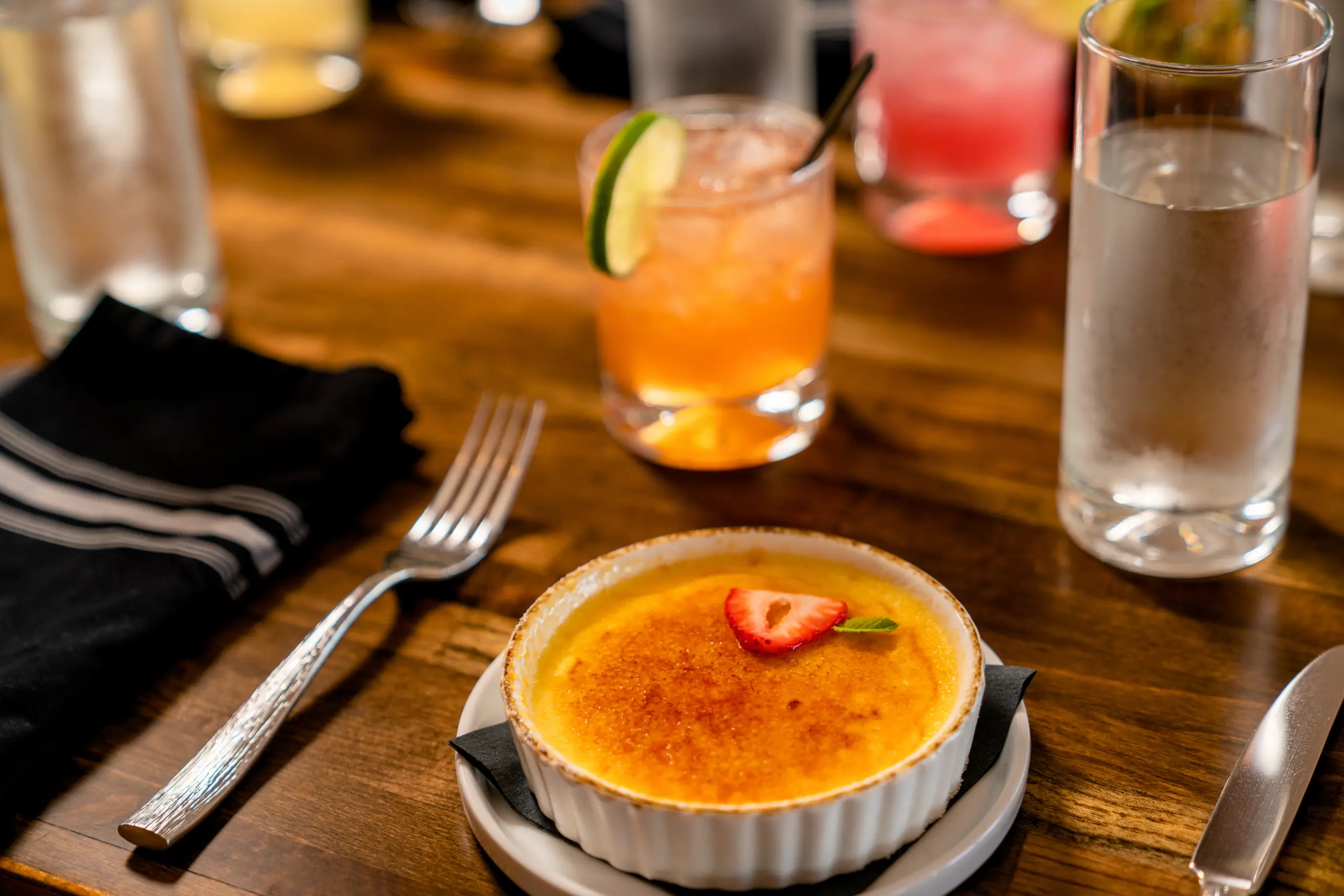 A ramekin of Crème Brûlée, garnished with a strawberry and mint, sits next to silverware on a wooden table. An orange cocktail and water glass are in the background.