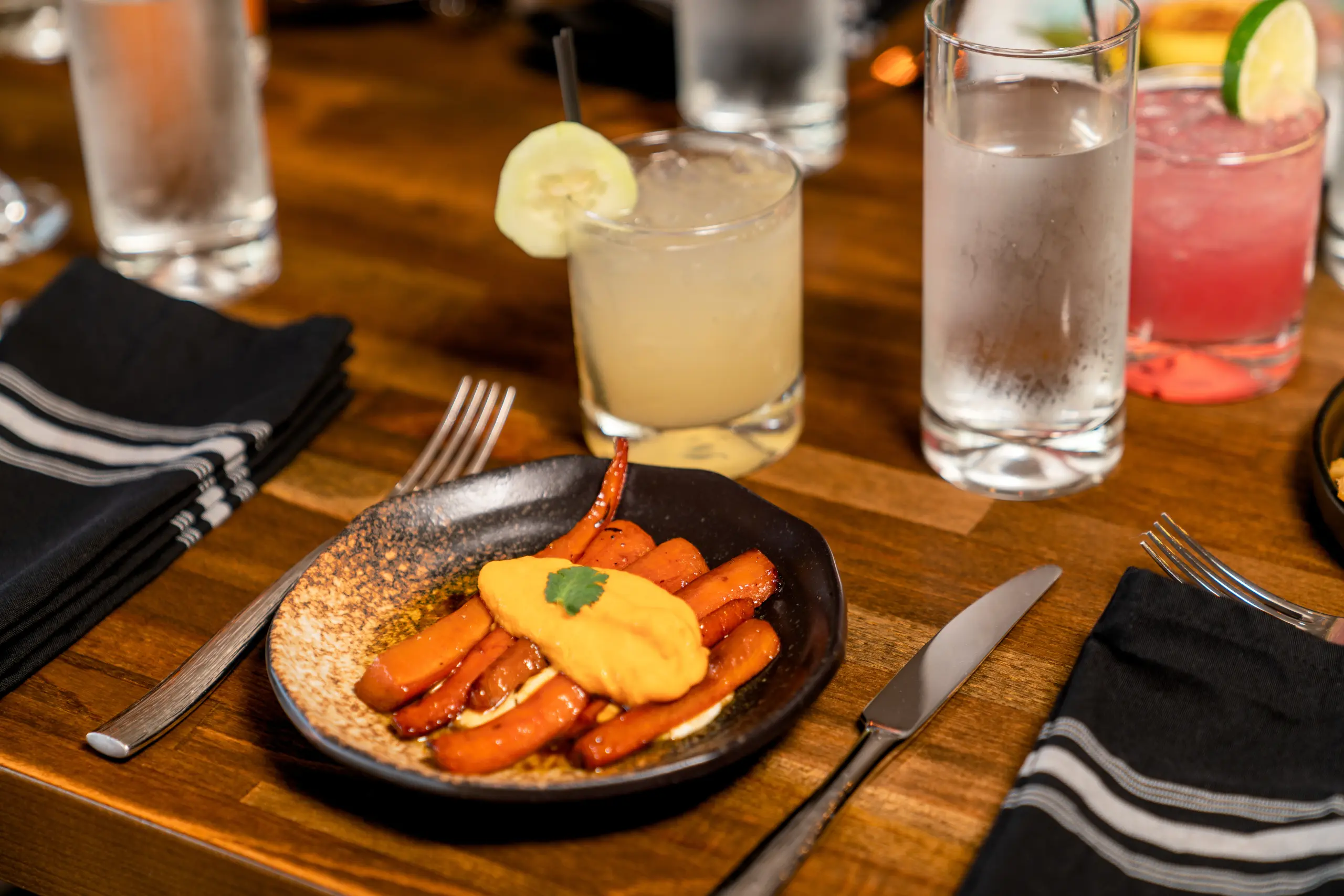 A dark plate of glazed carrots with an orange puree sits on a wooden restaurant table, flanked by silverware and black napkins. Glasses of water and two cocktails, one with cucumber and one with lime, are nearby.