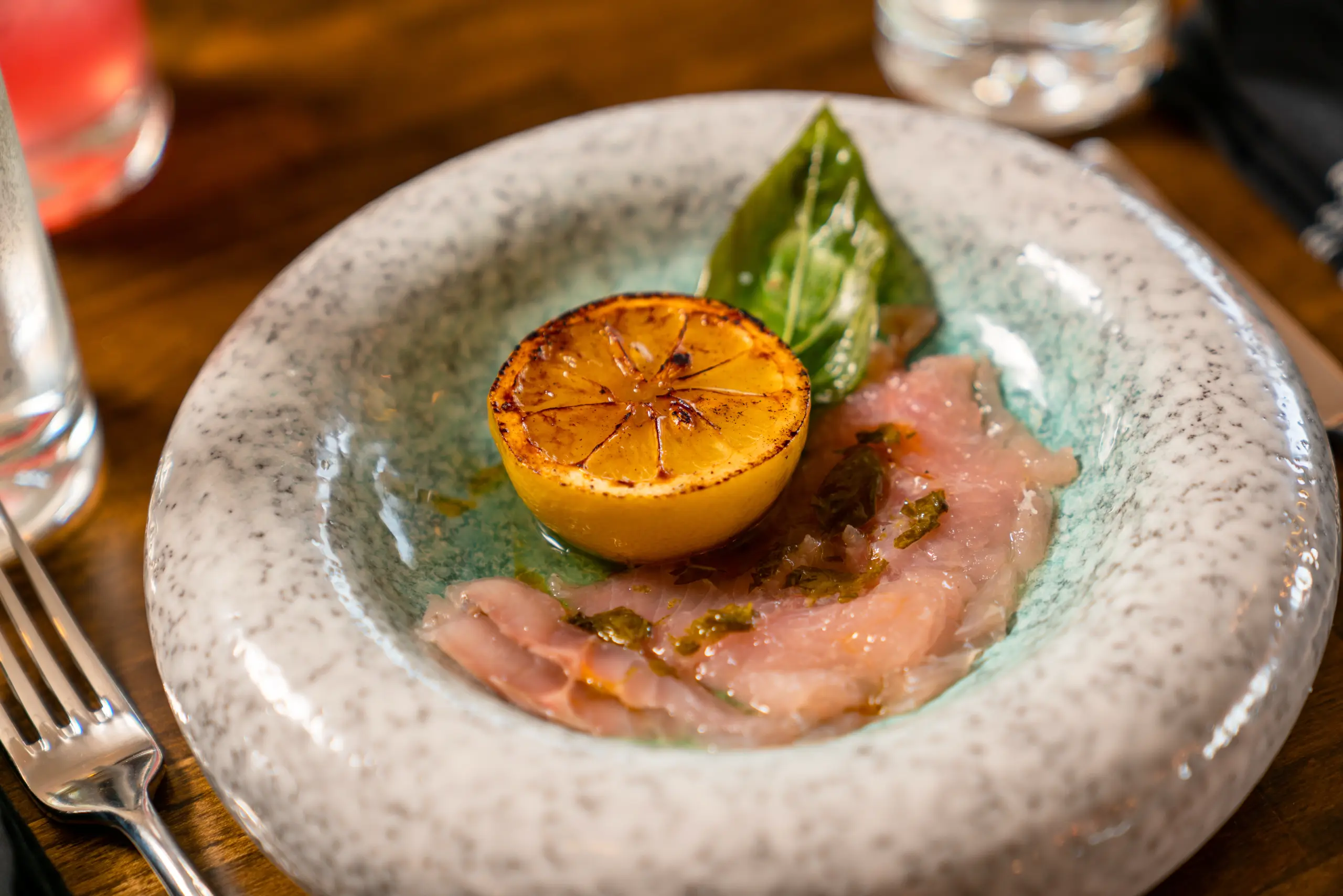 A restaurant dish of Great Lakes Crudo, featuring thinly sliced fish topped with green oil and a basil leaf, next to a heavily grilled lemon half.