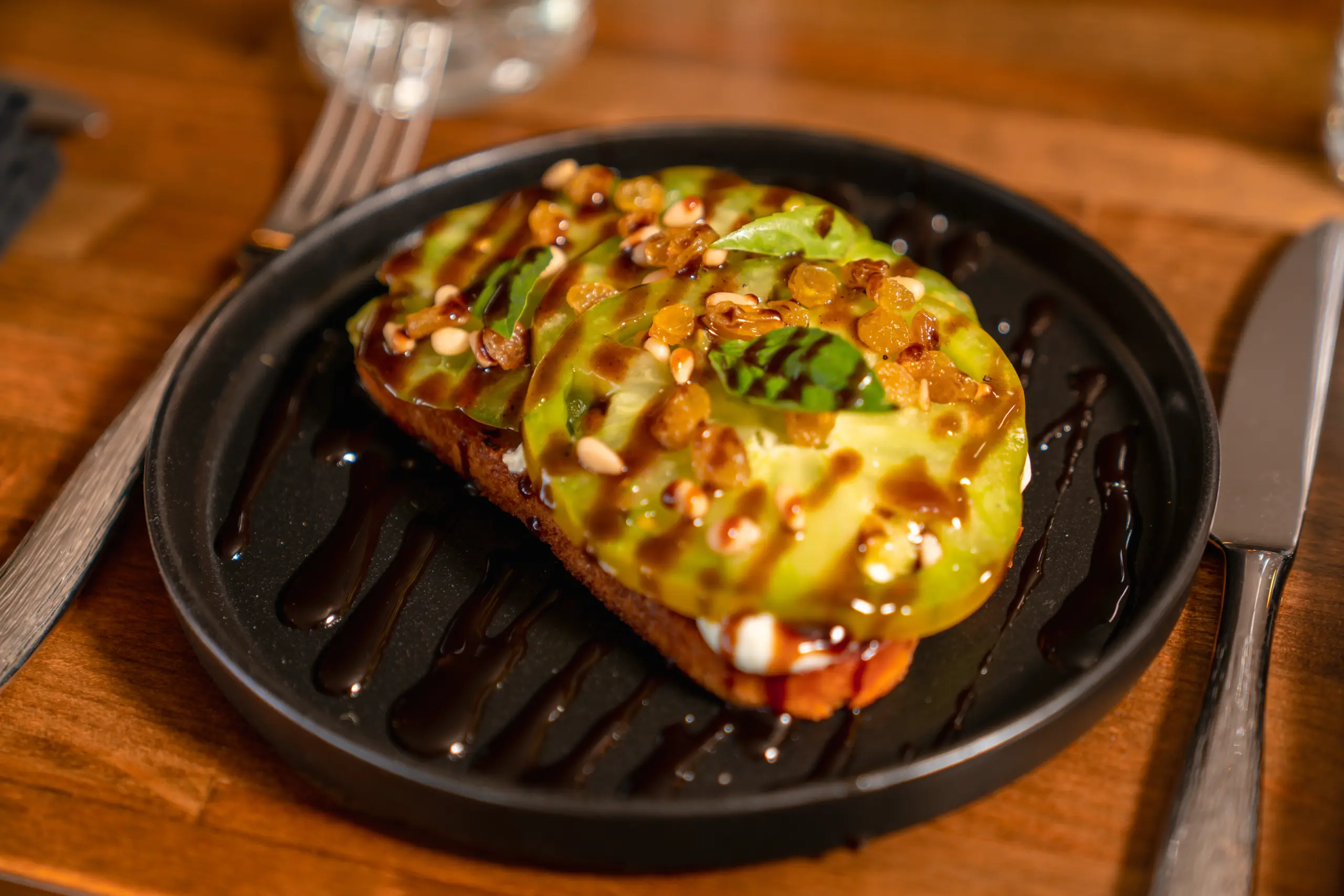 An appetizing heirloom tomato and ricotta tartine, covered in pine nuts, golden raisins, and a dark balsamic reduction, served on a black plate.