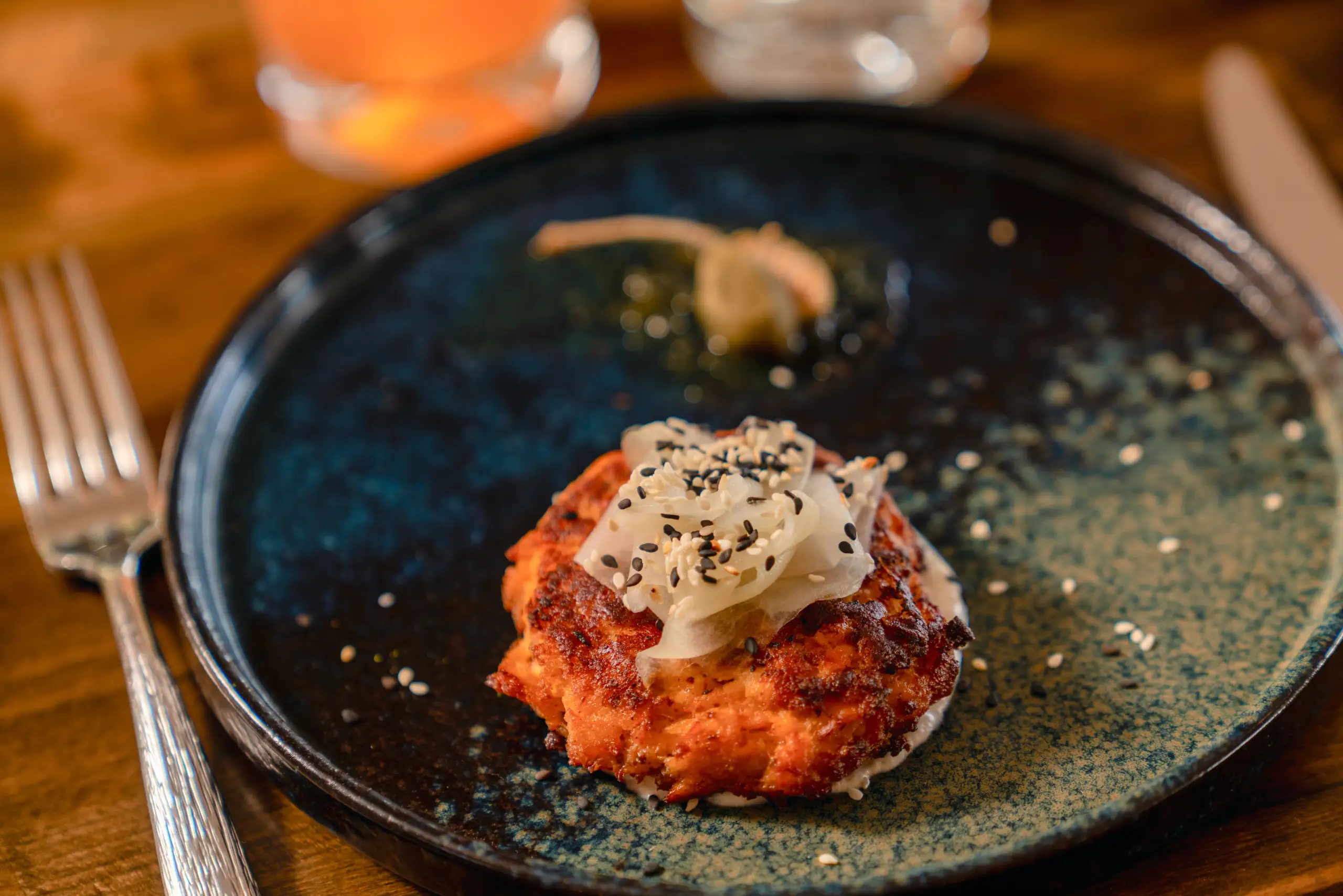 A single salmon cake topped with pickled vegetables and sesame seeds is served on a speckled blue ceramic plate next to a fork.