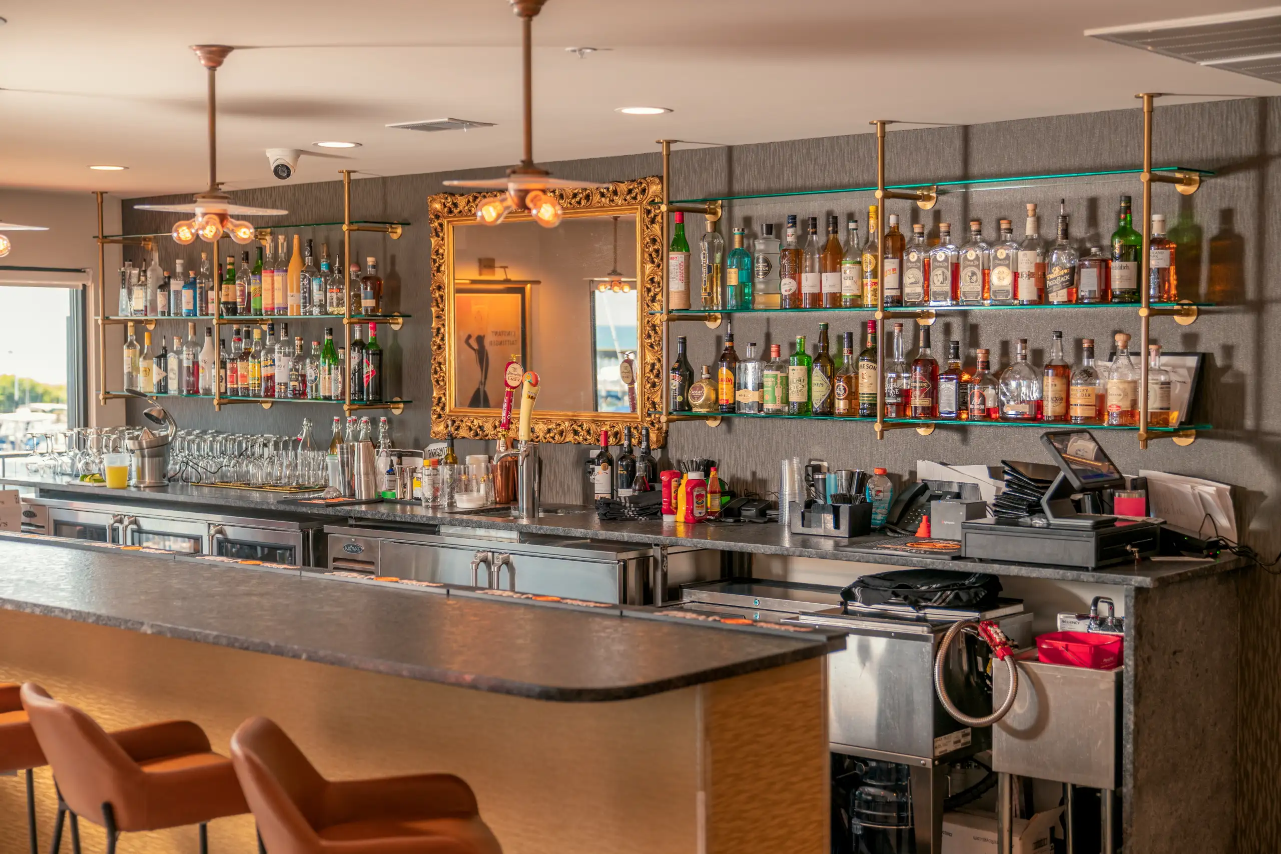 A well-equipped restaurant bar with a dark stone countertop and three orange leather bar stools in the foreground. Glass shelves displaying liquor bottles flank an ornate gold mirror.