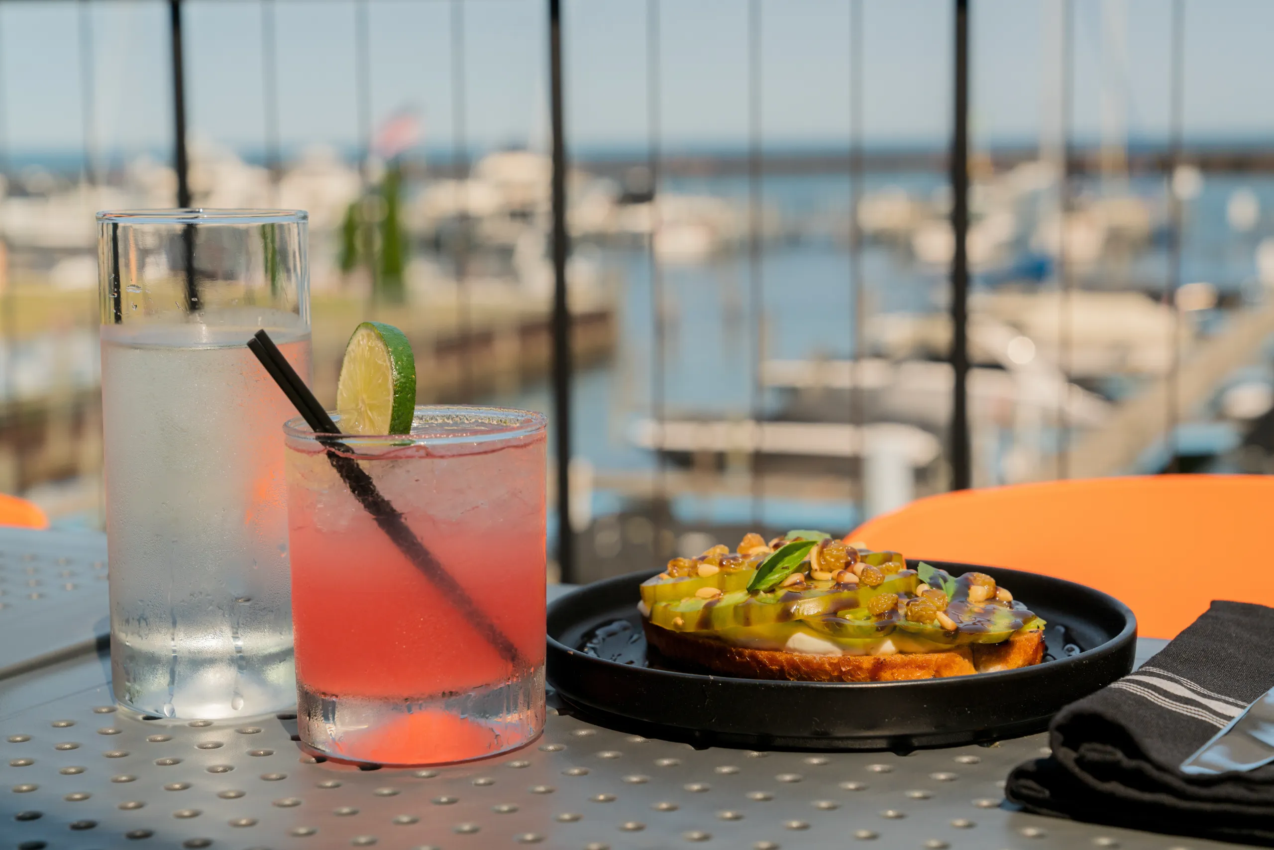 A refreshing pink cocktail and chilled water are on a table next to toast topped with green tomatoes and nuts, with a bright, sunny marina background.
