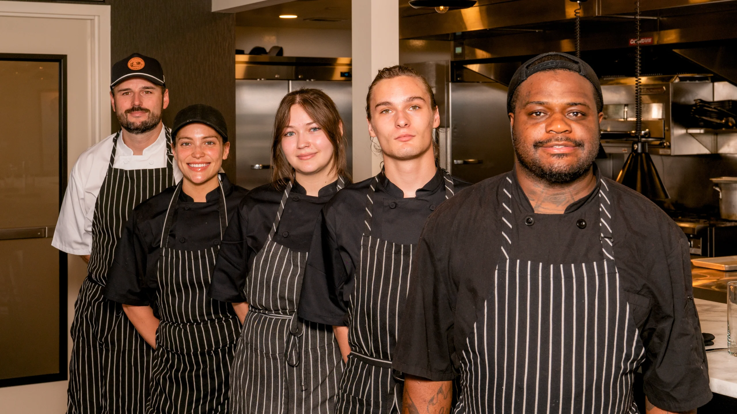 A diverse team of five kitchen staff members, all wearing pinstripe aprons, smile confidently while posing in the restaurant kitchen.