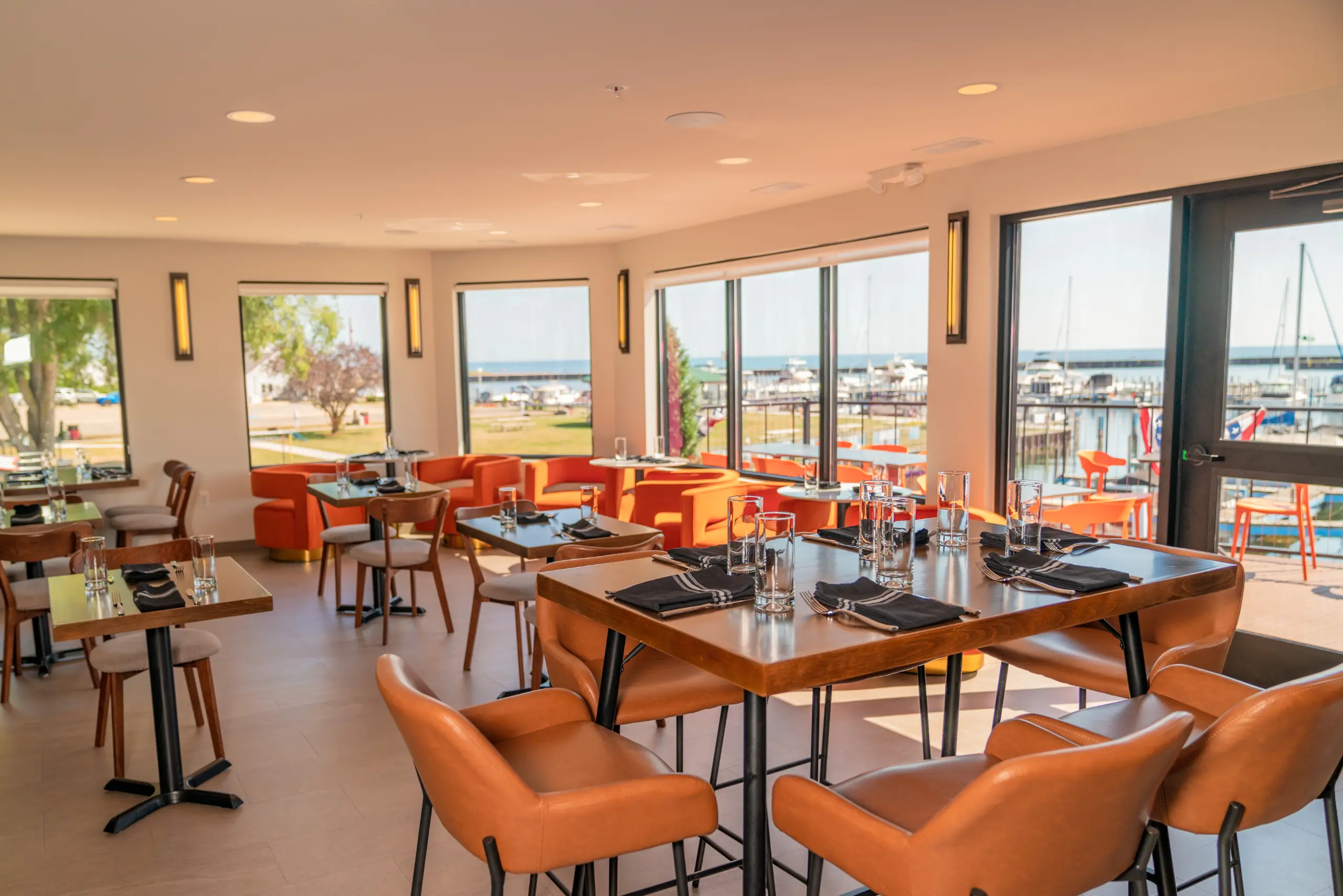 A bright restaurant dining room overlooking a marina, featuring wooden tables set with glasses and black napkins. The seating is a mix of orange lounge chairs and brown leather dining chairs.