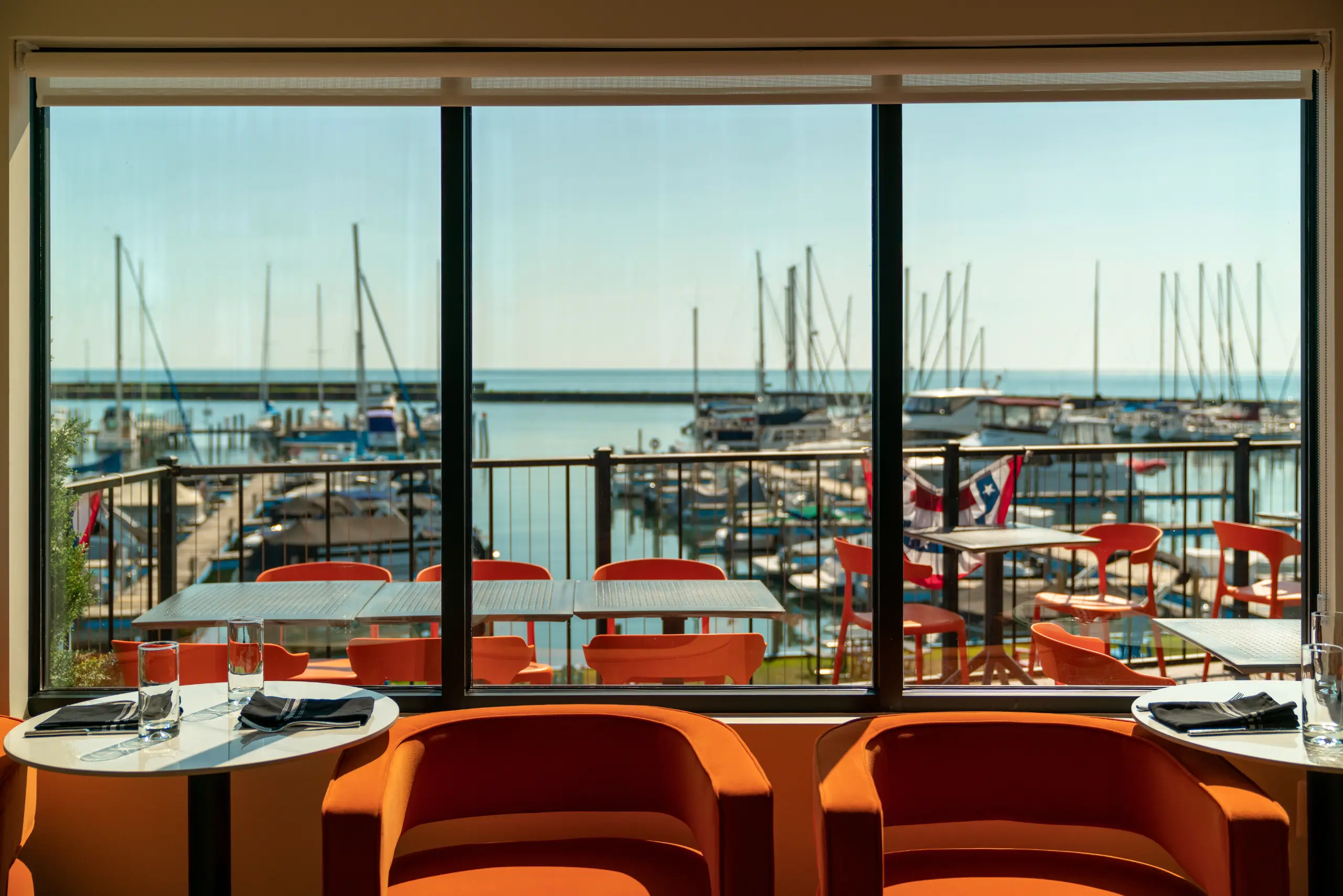 Restaurant seating in vibrant orange colors overlooks a marina view through large windows, featuring boats docked in bright sunshine.