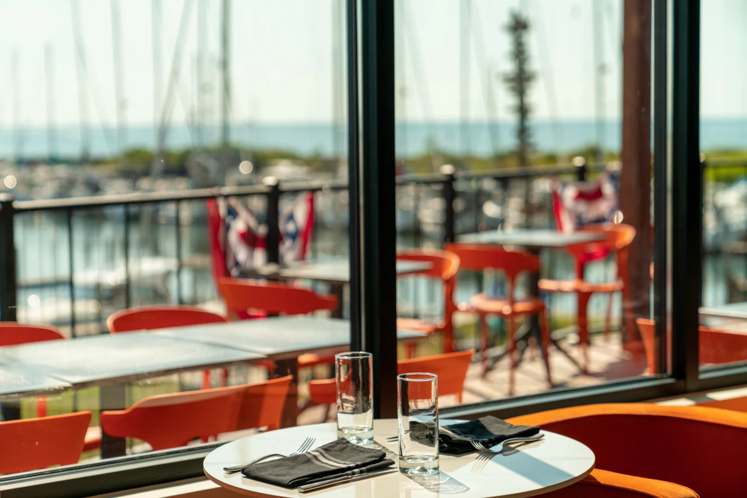 A white restaurant table with two water glasses and black napkins is set near a window overlooking a marina and bright orange outdoor seating.